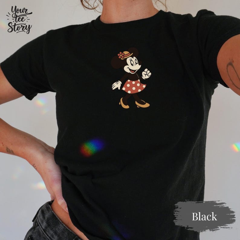 May include: Black t-shirt featuring a cartoon Minnie Mouse design. Minnie Mouse is wearing a red polka dot dress, a hat, and heels. The shirt has the text "Your Tee Story" on the upper left and "Black" on the lower right.
