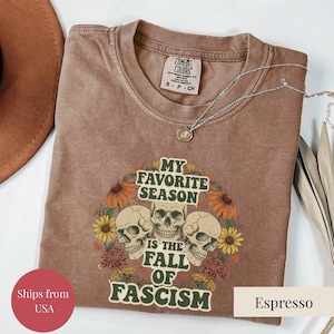 May include: Espresso-colored t-shirt with the text "MY FAVORITE SEASON IS THE FALL OF FASCISM" and a graphic of three skulls surrounded by flowers. A silver necklace is draped across the shirt.
