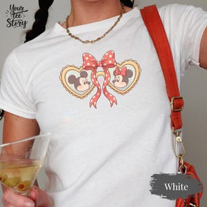 Op de afbeelding: Wit T-shirt met een Mickey en Minnie Mouse ontwerp. Het ontwerp bevat twee hartvormige frames met Mickey en Minnie, verbonden door een roze strik met witte stippen. Het shirt heeft de tekst "Your Tee Story" linksboven.