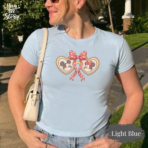 Op de afbeelding: Lichtblauw T-shirt met een afbeelding van Mickey en Minnie Mouse in hartvormige kaders, verbonden door een rode strik met witte stippen. Het shirt heeft korte mouwen en de tekst "Your Tee Story" is zichtbaar.