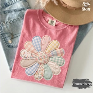 以下が含まれることがあります： ピンク色のComfort ColorsのTシャツ。大きなアップリケのデイジーのデザインが特徴です。 デイジーの花びらは、花柄、ギンガム、チェック柄など、さまざまな柄の生地で作られています。 青いジーンズとタン色の帽子と一緒に表示されています。「Your Tee Story」と「Churnchberry」の文字が見えます。