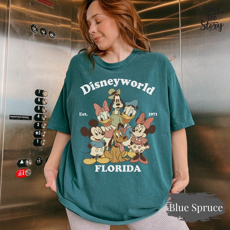 May include: Blue spruce-colored t-shirt with a Disneyworld graphic featuring Mickey and Minnie Mouse, Donald and Daisy Duck, Goofy, and Pluto. The text "Disneyworld Florida Est. 1971" is printed on the front. The shirt is a comfortable, oversized fit.