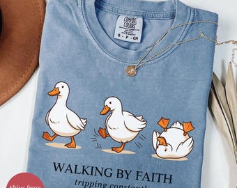 May include: Blue jean colored t-shirt with a graphic of three white ducks, one walking, one tripping, and one upside down. The text "WALKING BY FAITH tripping constantly" is below the ducks. A gold necklace is draped across the shirt.