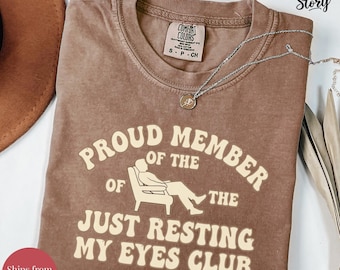 May include: Espresso-colored t-shirt with the text "PROUD MEMBER OF THE JUST RESTING MY EYES CLUB" in a retro font. The shirt features a graphic of a person reclining in a chair. A silver necklace is draped across the shirt.