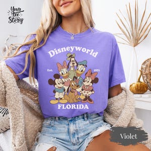 May include: Violet-colored t-shirt featuring a Disney World graphic. The design includes Mickey Mouse, Minnie Mouse, Donald Duck, Daisy Duck, Goofy, and Pluto. The text "Disneyworld Florida" is printed above the characters, with "Est. 1971" below.