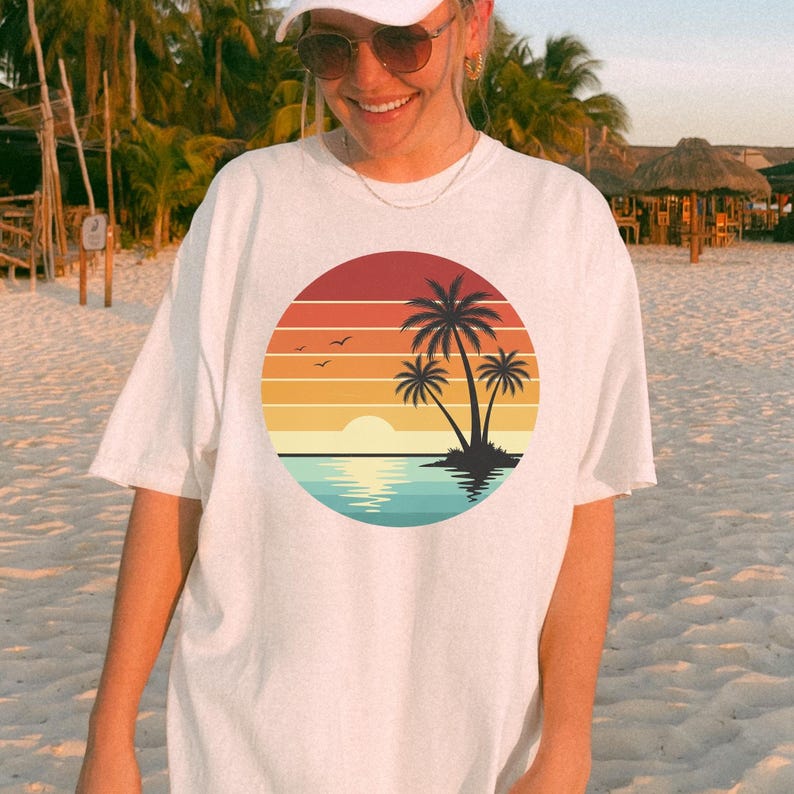 May include: White t-shirt featuring a circular graphic of a sunset over the ocean with palm trees. The design includes a gradient of orange, yellow, and blue hues, with a silhouette of palm trees and birds. The shirt is a casual, oversized fit.