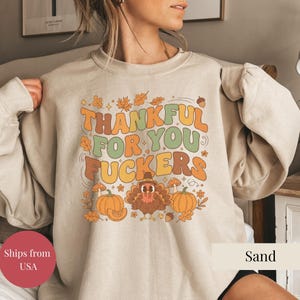以下が含まれることがあります： オレンジ、緑、茶色で「THANKFUL FOR YOU FUCKERS」と書かれたベージュのスウェットシャツ。デザインには、秋の葉、カボチャ、キノコ、巡礼者の帽子をかぶった七面鳥が含まれています。「Sand」の文字が右下に印刷されています。