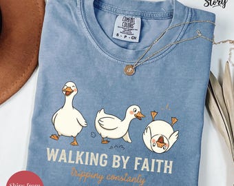 May include: Blue jean Comfort Colors t-shirt with a graphic of three cartoon ducks. The ducks are in various poses, with the text "WALKING BY FAITH tripping constantly" below. A gold necklace is draped across the shirt.