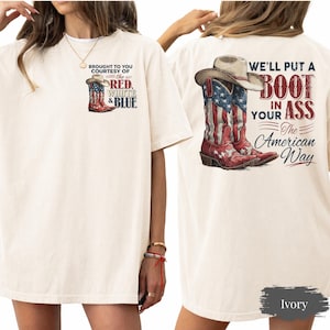 May include: Ivory-colored t-shirt featuring a graphic of cowboy boots with the American flag design and the text "BROUGHT TO YOU COURTESY OF THE RED, WHITE & BLUE." The back of the shirt displays a cowboy hat and boots with the text "WE'LL PUT A BOOT IN YOUR ASS The American Way."