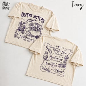 May include: An ivory t-shirt featuring dark purple ink graphics. The front displays a mortar and pestle with the text "OWENS SISTERS APOTHECARY EST. 1995". The back features botanical illustrations and the text "Always throw SPILLED SALT over your left shoulder, Keep ROSEMARY by your Garden Gate, Plant LAVENDER for luck, and fall in LOVE whenever you can."