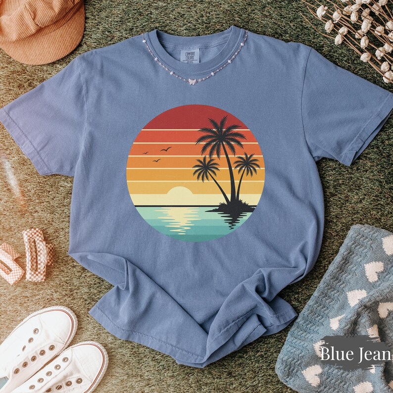 May include: A blue t-shirt featuring a sunset design with palm trees, a sun, and water. The graphic is a circle with horizontal stripes of red, orange, yellow, and teal. The shirt is surrounded by accessories.
