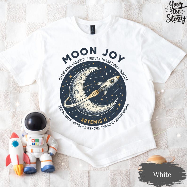 May include: White t-shirt with a graphic design featuring a crescent moon, a rocket, and the text "MOON JOY" and "ARTEMIS II". The design celebrates humanity's return to the lunar frontier. The names of the astronauts are also listed.