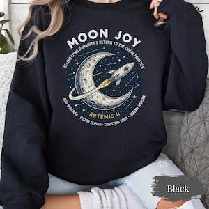 May include: Black sweatshirt with a graphic featuring a crescent moon, a rocket, and the text "MOON JOY" and "ARTEMIS II". The design celebrates humanity's return to the lunar frontier. Crew names are also listed.
