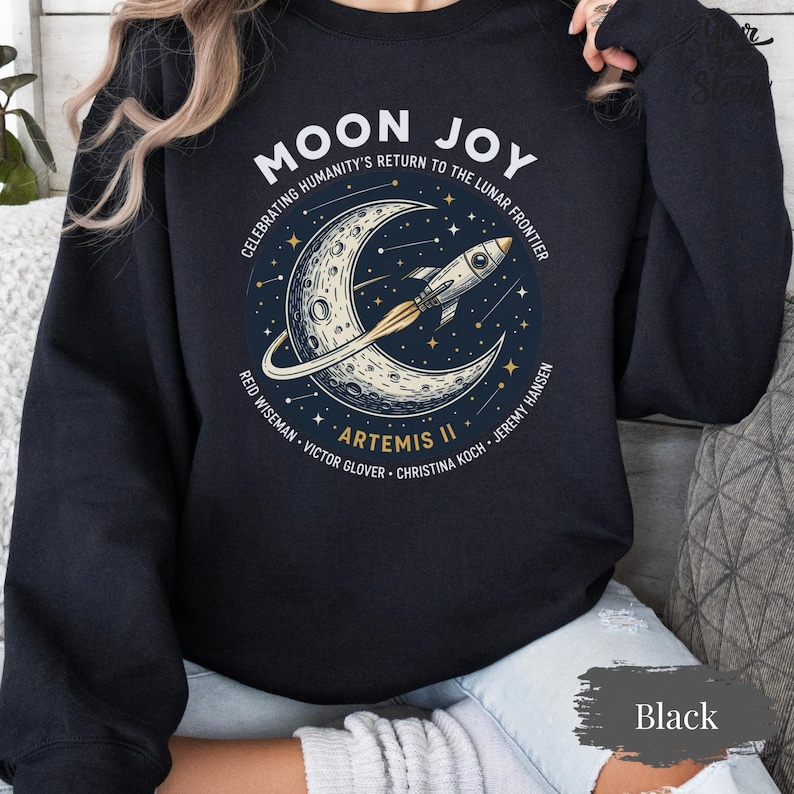 May include: Black sweatshirt with a graphic featuring a crescent moon, a rocket, and the text "MOON JOY" and "ARTEMIS II". The design celebrates humanity's return to the lunar frontier. Crew names are also listed.