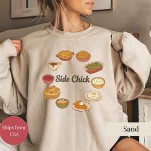 May include: Sand-colored sweatshirt with a circular graphic of Thanksgiving side dishes, including mac and cheese, stuffing, and mashed potatoes. The words "Side Chick" are in the center of the design. The sweatshirt is a neutral color.