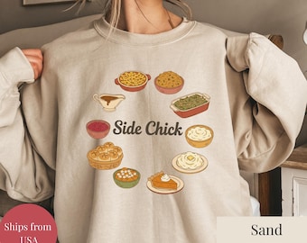 May include: Sand-colored sweatshirt with a circular graphic of Thanksgiving side dishes, including mac and cheese, stuffing, and mashed potatoes. The words "Side Chick" are in the center of the design. The sweatshirt is a neutral color.