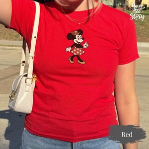 May include: A red t-shirt featuring a cartoon Minnie Mouse design. Minnie Mouse is wearing a brown hat, a polka-dot skirt, and brown shoes. The shirt also has the text "Your Tee Story" and "Red".
