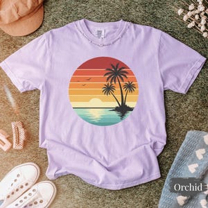 May include: A light purple t-shirt featuring a sunset design with palm trees, a sun, and water. The graphic is inside a circle. The shirt has a distressed neckline and short sleeves. The word "Orchid" is on a nearby blanket.