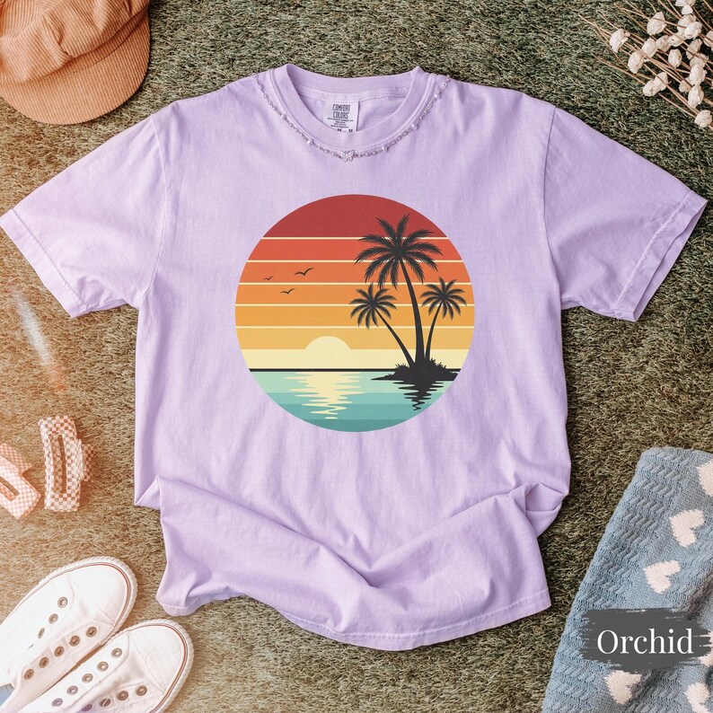 May include: A light purple t-shirt featuring a sunset design with palm trees, a sun, and water. The graphic is inside a circle. The shirt has a distressed neckline and short sleeves. The word "Orchid" is on a nearby blanket.