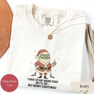 Puede incluir: Camiseta de color marfil con un diseño de rana navideña. La rana lleva un traje de Papá Noel y sostiene una taza. El texto dice: "I HAVE TO SAY WEIRD STUFF OR I'LL DIE - BUT MERRY CHRISTMAS!" Un collar dorado está sobre la camiseta.