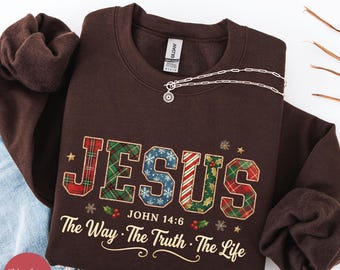 Op de afbeelding: Donker chocoladekleurige sweatshirt met het woord "JESUS" in een ruit- en sneeuwvlokontwerp. De tekst "JOHN 14:6" en "The Way. The Truth. The Life" staan eronder. Een zilveren ketting met een hanger ligt op de sweatshirt.