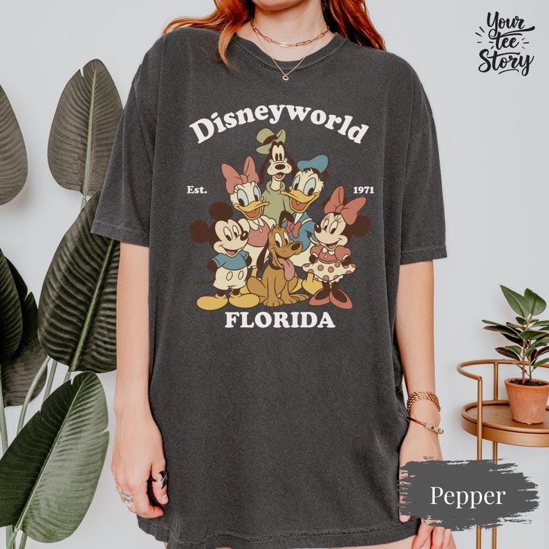 May include: Dark gray t-shirt featuring a vintage-style graphic of Disney characters, including Mickey and Minnie Mouse, Donald and Daisy Duck, and Goofy. The text "Disneyworld Florida Est. 1971" is printed above and below the characters.