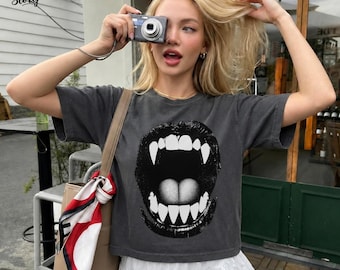 May include: A charcoal gray short-sleeve t-shirt featuring a large graphic print of an open mouth with sharp white fangs. The shirt has a crew neckline and a relaxed fit. A white, red, and black patterned scarf is attached to a tan shoulder bag.