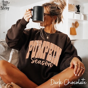 Puede incluir: Una sudadera de cuello redondo en color marrón chocolate oscuro con el texto PUMPKIN en una fuente grande de cuadros vichy naranja y blanco con pequeñas ilustraciones de lazos, y la palabra season debajo en una fuente cursiva clara.