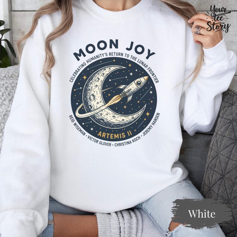 May include: White sweatshirt with a circular graphic featuring a crescent moon, a rocket, and the text "MOON JOY" and "ARTEMIS II". The graphic also includes the text "CELEBRATING HUMANITY'S RETURN TO THE LUNAR FRONTIER".