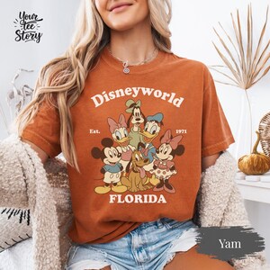 May include: A yam-colored t-shirt featuring a retro Disney World design with Mickey Mouse, Minnie Mouse, Donald Duck, Daisy Duck, Goofy, and Pluto. The text "Disneyworld Florida Est. 1971" is printed in a vintage style.