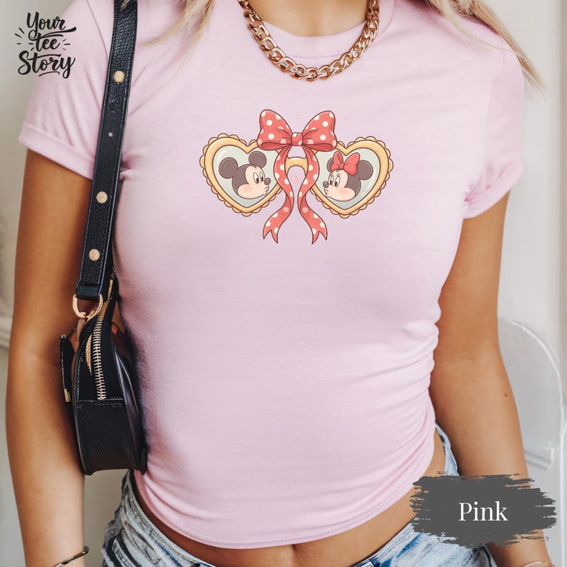Op de afbeelding: Zachtroze T-shirt met een afbeelding van Mickey en Minnie Mouse in hartvormige kaders, verbonden door een rode strik met stippen. Het shirt heeft korte mouwen en de tekst "Your Tee Story" is linksboven zichtbaar.