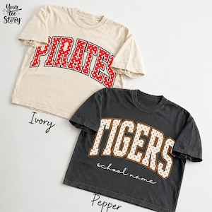 May include: Two short-sleeve crew neck t-shirts. The ivory shirt features the red text PIRATES with a white polka dot pattern. The pepper gray shirt features the orange text TIGERS with a white polka dot pattern and the text school name below.