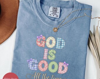 Puede incluir: Camiseta azul vaquero con la frase "GOD IS GOOD All the time" en letras de patchwork coloridas. Un collar de oro con un colgante descansa sobre la camisa. La camisa está doblada y se muestra sobre una superficie blanca.