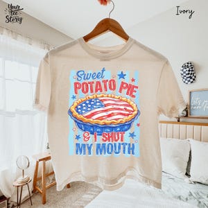 Op de afbeelding: Een ivoorkleurig t-shirt met korte mouwen en een afbeelding van een taart met een vulling in het patroon van de Amerikaanse vlag. De tekst Sweet POTATO PIE & I SHUT MY MOUTH staat in rood en blauw, omringd door lichtblauwe strepen en sterren.