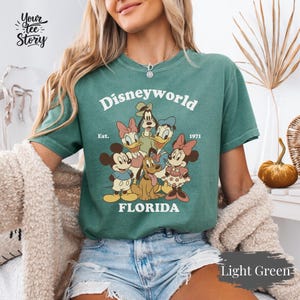 May include: Light green t-shirt featuring a graphic of Disney characters, including Mickey and Minnie Mouse, Donald and Daisy Duck, Goofy, and Pluto. The text "Disneyworld FLORIDA" is printed above the characters, with "Est. 1971" below. The shirt is a light green color.