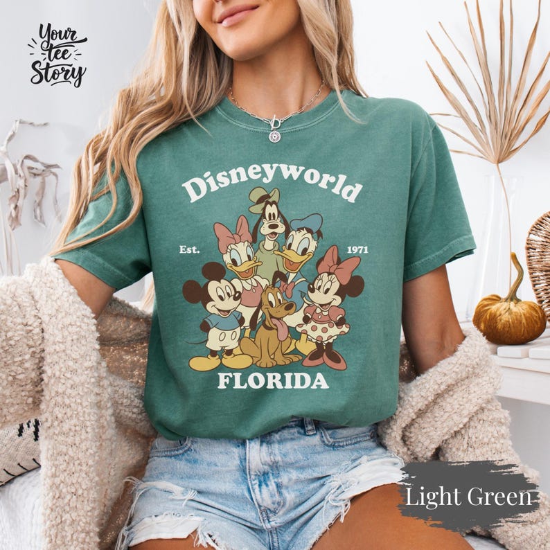 May include: Light green t-shirt featuring a graphic of Disney characters, including Mickey and Minnie Mouse, Donald and Daisy Duck, Goofy, and Pluto. The text "Disneyworld FLORIDA" is printed above the characters, with "Est. 1971" below. The shirt is a light green color.