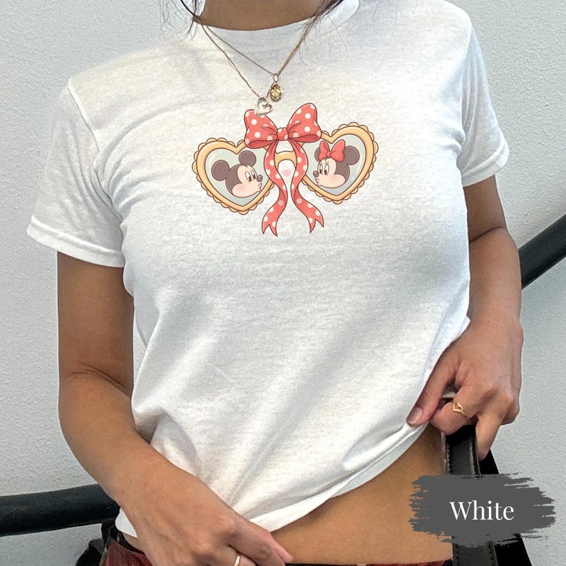 Op de afbeelding: Wit T-shirt met een Mickey en Minnie Mouse ontwerp. De afbeelding bevat hartvormige kaders met de gezichten van de personages, een rode strik met stippen en linten. Het T-shirt heeft korte mouwen en is gemaakt van zacht materiaal.