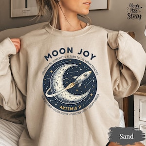May include: Sand-colored sweatshirt with a circular graphic featuring a crescent moon, stars, and a rocket ship. Text includes "MOON JOY" and "ARTEMIS II", celebrating humanity's return to the lunar frontier. The sweatshirt is a neutral color.