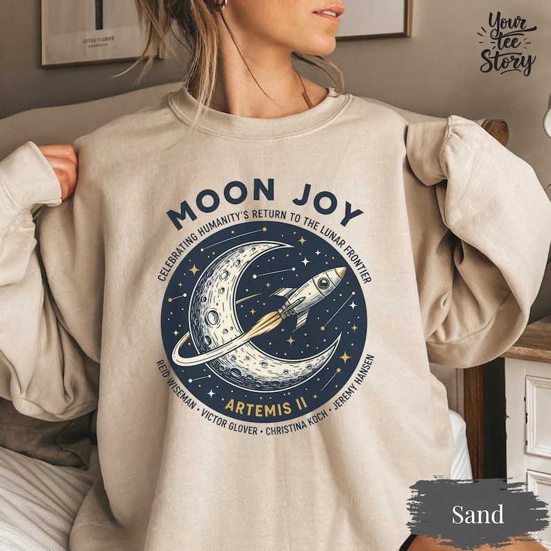 May include: Sand-colored sweatshirt with a circular graphic featuring a crescent moon, stars, and a rocket ship. Text includes "MOON JOY" and "ARTEMIS II", celebrating humanity's return to the lunar frontier. The sweatshirt is a neutral color.