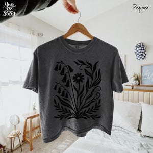 May include: A dark gray t-shirt with a black floral design, including bell-shaped flowers and a daisy. The shirt is hanging on a wooden hanger. The text "Your Tee Story" is visible on the upper left. The word "Pepper" is written on the upper right.