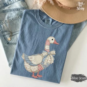 Könnte beinhalten: Blaues Comfort Colors T-Shirt mit einem Patchwork-Gänsemotiv. Die Gans hat einen roten Schnabel und Füße sowie eine blaue Schleife. Das Design beinhaltet florale, Vichy- und unifarbene Muster. Der Text "Blue Jean" ist unten rechts aufgedruckt.