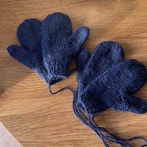 Könnte beinhalten: Zwei Paar marineblaue Strickfäustlinge werden auf einer Holzoberfläche gezeigt. Die Fäustlinge sind handgefertigt, mit sichtbaren Nähten und einem strukturierten Aussehen. Jeder Fäustling hat eine Schnur zum Aufhängen oder Binden. Die Fäustlinge sind für Wärme und Schutz konzipiert.