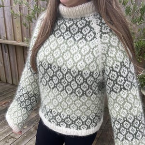 以下が含まれることがあります： ハイネックカラーの、手編みのセーター。白、ライトグリーン、ダークグレーの色合いで、繰り返しのひし形模様が特徴です。袖と襟は無地のオフホワイトです。