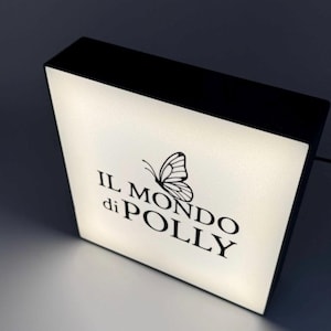 May include: A square, illuminated sign with a black frame. The sign's face is white with the text "IL MONDO di POLLY" and a butterfly graphic in black. The sign is lit from within, creating a soft glow.