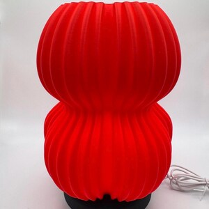 Peut inclure: Lampe de table rouge vif, côtelée, avec un design empilé et arrondi. La lampe repose sur une base noire et possède un cordon d'alimentation blanc. La forme et la couleur vives de la lampe en font un élément décoratif saisissant.