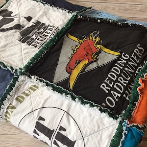 Op de afbeelding: Een patchworkdeken gemaakt van hergebruikte T-shirts. De deken heeft een verscheidenheid aan kleuren en ontwerpen, waaronder een rode en gele vogel, een groen en wit ontwerp en een blauw en wit ontwerp. De deken is gemaakt met een rafelige rand.