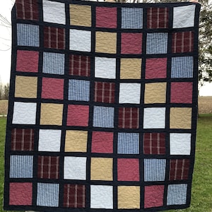 Op de afbeelding: Een quilt gemaakt van vierkanten van verschillende stoffen in tinten blauw, bruin, wit en rood. De vierkanten zijn aan elkaar gestikt met een donkerblauwe stof en een zwarte rand.