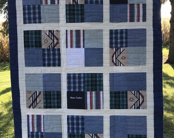 May include: A patchwork quilt featuring a grid of squares made from various blue, green, and tan fabrics, including plaid, denim, and patterned textiles. The quilt has a dark blue border and includes the text Roy and Alsum Produce on two squares.