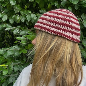 May include: A crochet beanie featuring horizontal stripes in alternating shades of deep red and light cream. The hat has a rounded crown and a simple, textured stitch pattern, designed to fit snugly around the head with a clean, finished edge.