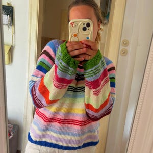 Könnte beinhalten: Ein langärmeliger Strickpullover mit horizontalen Streifen in einer lebendigen Palette aus Blau, Grün, Pink, Orange, Gelb und Weiß. Das Kleidungsstück hat einen gerippten Rundhalsausschnitt und gerippte Bündchen mit einem mehrfarbigen Garnmuster.